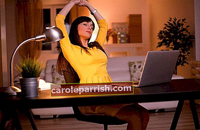 wanita berambut cokelat dengan gaun kuning dengan rambut panjang berdiri di depan komputernya di mejanya dengan lampu di depan sofa putih